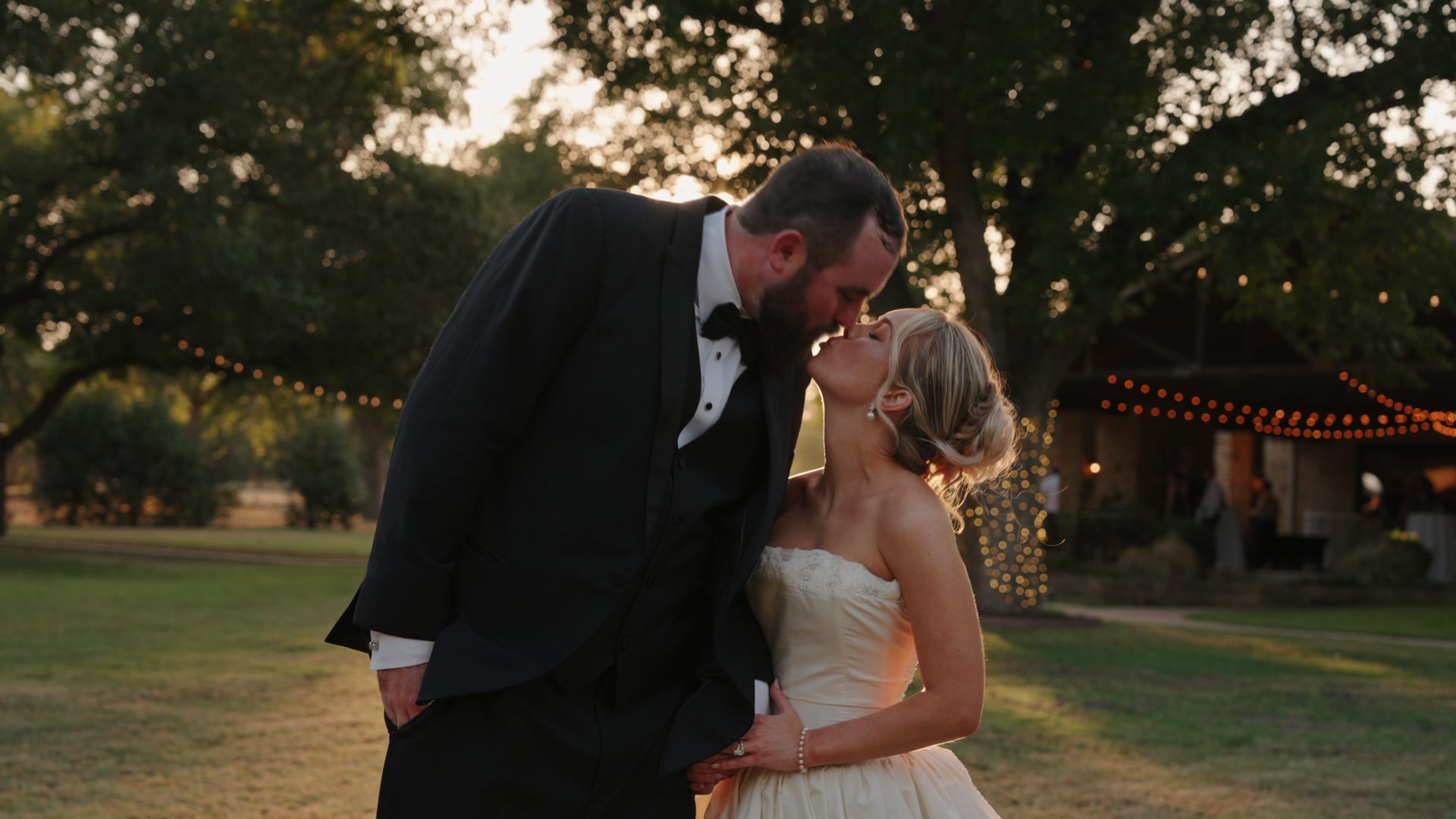A groom kissing his bride at golden hour under string lights at The Orchard